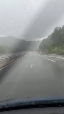 The video shows a first-person perspective from inside a vehicle driving on a wet road, likely a highway or major route, through a mountainous or hilly area. The weather is rainy and overcast, with numerous raindrops visible on the windshield, blurring the view slightly. The road surface is visibly wet, reflecting the gray sky.

On both sides of the road, guardrails are present. Hills and mountains, covered with dense green trees, recede into the misty background. The overall atmosphere is somber due to the rain and low clouds.

As the vehicle progresses, several traffic cones with blue and white chevron patterns, indicating a curve or lane direction, become visible on the right side of the road. Further ahead, circular blue road signs with white arrows, also directing traffic, can be seen. The road appears to curve gently to the right in the distance. No people or other vehicles are clearly visible, and no discernible text is present on any signs, only symbolic arrows. The scene suggests driving conditions during a rainy day in a natural, rugged landscape, consistent with the provided context of Dragoman, Bulgaria.