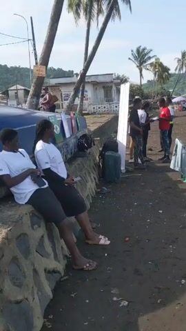 Here is a description of the video:

The video shows a group of people standing on a dark, sandy beach under a bright, sunny sky. In the foreground, two women are sitting on a low concrete wall, both looking at their phones. Behind them, there are some posters and luggage stacked against the wall. A group of people are gathered around a man in a red shirt and another in a safety vest who appears to be holding paperwork. Several other people are standing nearby, some holding clipboards, seemingly engaged in conversation. 

In the background, there are palm trees and buildings, and beyond that, a body of water with a visible coastline in the distance. The beach appears to have a significant amount of debris scattered around. Several wheelbarrows can also be seen nearby, adding a touch of utilitarian equipment to the beach scene.