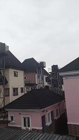 The video shows a collection of houses with dark-tiled roofs and light-colored walls. Several of the houses have tall, metal water towers supporting black water tanks on their roofs. The towers have ladders and railings. The sky is overcast.