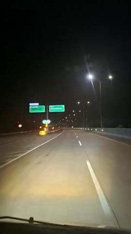 A forward-facing dashboard camera shot shows a vehicle driving at night on a multi-lane highway. The highway is illuminated by numerous streetlights on tall poles. Two green road signs are visible on the left side of the road. The road has white lane markings and reflective markers along the edges. The vehicle's headlights illuminate the immediate road ahead in a yellow hue. There are no other vehicles visible in the frame, and the highway appears empty. The overall scene is dark, with the highway and its immediate surroundings being the primary source of light.