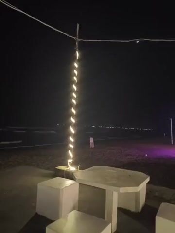 The video shows a nighttime view of an outdoor seating area on what appears to be a beach or waterfront. In the foreground, there is a concrete table and several cube-shaped seating elements, all illuminated by white lights. A vertical pole wrapped in a string of yellow lights stands to the left. In the middle ground, there is a tall, thatched structure and two white spherical lights hanging from wires. Beyond this, the scene opens to a dark expanse, likely the sea, with faint lights visible in the distance. The background is dark, suggesting it is nighttime. Some purple lighting is present on the ground in the middle ground, illuminating some foliage and a small structure. The overall atmosphere is that of an outdoor cafe or lounge area at night.