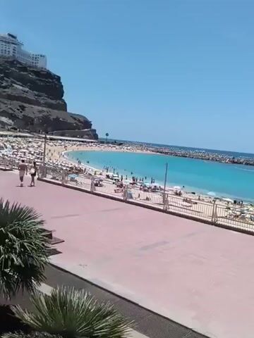 The video shows a sunny beach scene. The beach is crowded with people sunbathing, swimming, and walking along the shore. There are numerous sun umbrellas shading the sand. A rocky breakwater extends into the turquoise water, creating a calm area for swimming. In the background, a large, white hotel complex is built on top of a dark rocky cliff overlooking the beach. A tiled pedestrian walkway lined with a simple white fence runs along the top of the beach. A small palm tree is visible in the foreground.