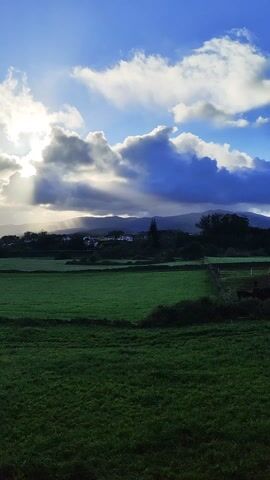 The scene shows a verdant field under a dynamic sky. The foreground is dominated by lush green grass, with some areas appearing slightly darker. In the midground, there are hints of houses or a small village nestled against a backdrop of distant hills. Above, the sky is a blend of deep blue and white, with fluffy clouds scattered throughout. Sunlight streams through the clouds, creating a dramatic effect of light and shadow. The overall impression is that of a serene and picturesque countryside landscape.