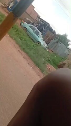 The video is filmed from the perspective of someone moving, likely in a vehicle, looking towards the right. In the foreground, a yellow metal bar with some rust and signs of welding is visible, partially obstructing the view. A dark-skinned knee or leg is also visible in the bottom right corner of the frame. The background shows a dirt road with some patches of grass and what appears to be a settlement. There are several buildings, some looking like simple houses and others resembling temporary structures or stalls made of corrugated metal. In the distance, a person in a light-colored suit is walking away from the camera across the road, near a puddle of water. There are also some containers and objects scattered around the area, suggesting a possibly unkempt environment. The overall lighting indicates it is daytime.