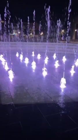 A nighttime outdoor scene shows a public plaza with a water fountain. The fountain consists of multiple jets of water, each illuminated by a colored light. The lights cycle through various colors, primarily shades of purple and pink, creating a vibrant display against the dark background. The plaza is paved with large square tiles. In the background, buildings with illuminated windows and trees are visible. The video captures the water jets intermittently turning on and off, with the lights intensifying and dimming in sync with the water flow.