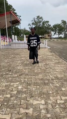 A young person with dark skin and hair styled in four buns walks towards the camera. They are wearing a black, oversized jersey with white lettering that says "PERRIPAYS" and the number "25" in large white numbers. The jersey has white stripes on the sleeves. They are also wearing loose, dark denim pants with a silver chain attached to the left side. The person is walking on a paved surface made of rectangular stones arranged in a pattern. To the left of the person, there are bleachers with red and white seating, and a red-roofed structure. In the background, there are trees and a cloudy sky. The person stops walking and looks directly at the camera, then turns their head slightly to the right.