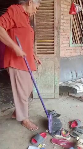 An elderly woman with grey hair is sweeping the floor with a purple broom. She is wearing an orange long-sleeved shirt and beige pants. Several pairs of sandals are scattered on the ground around her. The scene appears to be outdoors, near a wooden door with louvers and a brick wall.