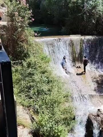 Two people are visible at the top of a cascading waterfall. One person, wearing a blue shirt and white shorts, is sliding down the wet rock face of the waterfall, while another person, wearing a dark shirt and black shorts, stands to the right, observing. Lush green foliage surrounds the waterfall and the rocky cliff face. The water flows down into a dark pool at the bottom.