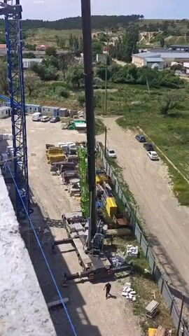 A tall blue construction crane is in the process of being assembled.  The main vertical tower of the crane is visible on the left. A long, horizontal blue boom section is being lifted by a separate, taller crane arm, which is positioned vertically in the center of the frame. This boom section is being attached to the top of the blue tower crane. Below the rising boom, construction materials, including stacks of concrete blocks and metal framing, are visible on the ground, along with temporary site offices and vehicles. The background consists of rolling green hills and some scattered buildings under a bright blue sky with scattered clouds.