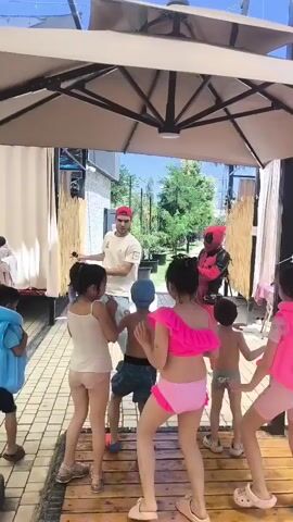 A group of children in swimwear are dancing outdoors under a large umbrella with a man dressed as Deadpool, a man in a bear costume, and a man wearing a red cap and a light-colored t-shirt, likely an entertainer or camp counselor. The children are positioned around the entertainers, some are facing away from the camera. The ground is paved with tiles, with a wooden platform in the center where some of the children are standing. In the background, there are white curtains, bamboo-like decorations, and greenery visible. The overall scene suggests a fun, outdoor event for children.
