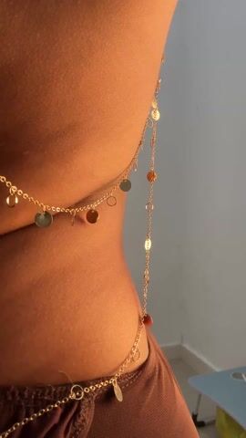 From behind, a woman wearing a brown dress with an open back stands still in front of a white wall. She is wearing a decorative gold body chain. The chain starts around her neck and descends to the waist and hip area. The dress has long, loose sleeves that hang down. The room is brightly lit. A corner of a blue-and-white table is visible in the bottom right corner.