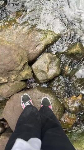 The video shows a first-person perspective of someone standing on rocks next to a shallow stream. The person is wearing black pants and black and pink sneakers. The stream has clear, flowing water with rocks visible on the streambed. The banks of the stream are lined with larger rocks and some greenery. The camera pans slightly to show more of the stream and the surrounding rocky terrain.