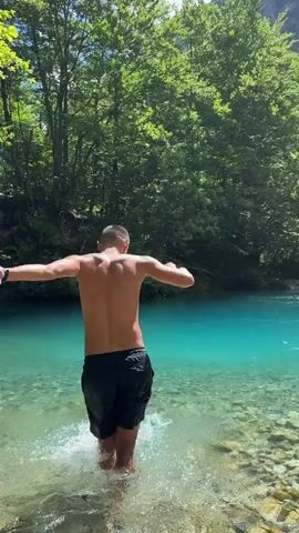 A young man, shirtless and wearing black shorts, stands waist-deep in a shallow, clear, turquoise river. He is facing away from the camera, with his arms extended slightly to his sides. The riverbed is covered in smooth, light-colored stones. Lush green trees with dense foliage line the background, creating a vibrant forest setting. Sunlight filters through the leaves, casting dappled shadows. The man is moving his arms in a playful manner, splashing the water. The camera angle is from behind the man, slightly elevated, capturing his upper body and the surrounding natural environment. The overall scene conveys a sense of refreshing immersion in nature on a bright, sunny day.