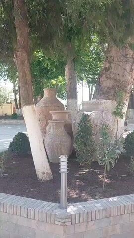The video shows a street side decoration made up of several carved stone urns arranged around the base of three trees inside a brick-walled planter. The trees are providing shade to the urns, which are decorated with various circular patterns.  A small cylindrical silver outdoor light is in the foreground.  The tree trunks have a white strip painted at the bottom.  A road and trees are visible in the background.