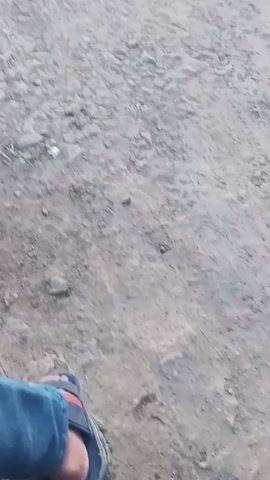 The video shows a dirt path with puddles of water and mud. There are several motorcycles parked on the side of the path, along with a few people. In the background, there are some buildings and trees. The sky is overcast, suggesting it may have recently rained or is about to rain. The overall scene appears to be in a rural or semi-rural setting.