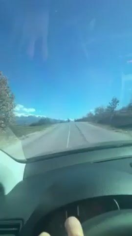 The video shows a car driving on a paved road, with snow-capped mountains in the distance under a bright blue sky with a few clouds. The road has a single white line marking the center. There are trees and shrubs on either side of the road. The dashboard of the car is visible, and a hand briefly appears to adjust something.