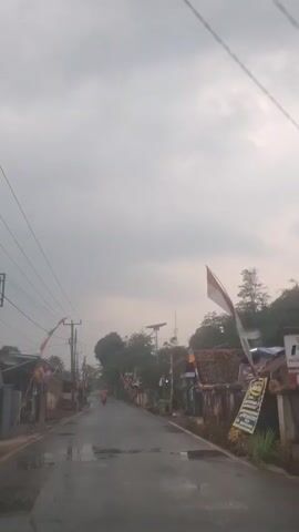 The video shows a road with houses on either side. There are Indonesian flags visible. The road is wet, suggesting it may have rained recently. A motorcycle with two people on it is traveling down the road. The sky is overcast.