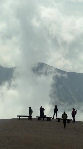 Six people are standing on a gravelly overlook in front of a mountain range partially obscured by clouds. Three of the people are standing near benches. One person is in the foreground, flying past from left to right, its wings spread. The sky is overcast with heavy clouds.