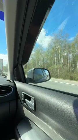 A view from inside a moving car, looking out the passenger-side window. The car is traveling on a paved road alongside a wooded area with birch trees. The sky is blue with scattered clouds. A large truck is visible in the distance. The car's side mirror reflects a blurred image of vehicles behind. The car's interior features a black textured door panel, a window switch, a door handle, and a portion of the dashboard with an air vent.  The car seems to be traveling at a moderate speed.