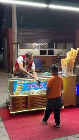 A man in a red vest and white shirt stands behind an ice cream cart and is interacting with a young boy in an orange shirt. The ice cream cart has colorful graphics of ice cream and the words "KAHRAMANMARAS DONDURMA" and "MEDO" illuminated on it. There are stacks of ice cream cones on a stand next to the cart. The man is holding a utensil and appears to be serving or preparing ice cream for the boy. The scene is taking place outdoors at night, with artificial lighting illuminating the area. There are other people in the background and a dark building behind the cart.