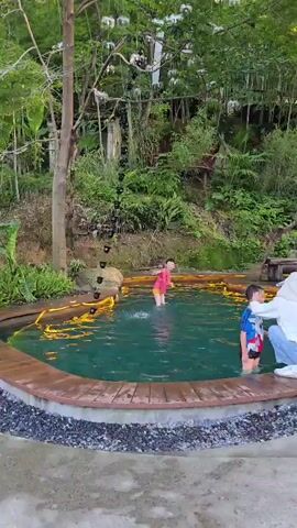 In this video, two young children are playing in a shallow, clear blue pool. The pool has a wooden deck surrounding it, with some rocks and lush green foliage creating a natural, outdoor setting. In the background, a person is seated on a wooden bench, observing the children. The scene appears to be a serene and playful environment. The lighting suggests it is daytime, possibly late afternoon, as the sun casts a warm glow. The presence of trees and dense vegetation hints at a location with abundant nature, consistent with the provided context of Xiaoshan District, China. The children seem to be enjoying themselves, splashing and moving around in the water, indicating a joyful and relaxed atmosphere. There is no visible text in the video.