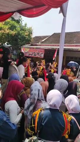 The video captures an outdoor cultural performance or parade, likely in an Asian country given the flags and attire. A crowd of people, many wearing headscarves and traditional clothing, are gathered under a red and white tent or awning. In the background, performers in elaborate costumes, including golden headdresses and ornate garments, are dancing. Two Indonesian flags are visible, one on a pole and another attached to a building in the background. The overall atmosphere appears festive and celebratory.