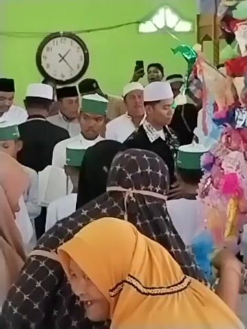 The video shows a gathering of people, primarily men wearing Muslim attire, in an indoor setting. The background includes a large clock on the wall and a brightly decorated structure adorned with colorful paper and fabric. In the foreground, a woman in a yellow hijab is looking towards the camera with an open mouth. To her right, another woman in a patterned hijab is reaching out towards the decorated structure. Several men are visible in the background, some holding phones, possibly recording the event. One man in the center appears to be speaking into a microphone, and a young man in a green skullcap is looking in his direction. The overall atmosphere seems to be festive or celebratory, with the decorated structure likely being a focal point of the event.