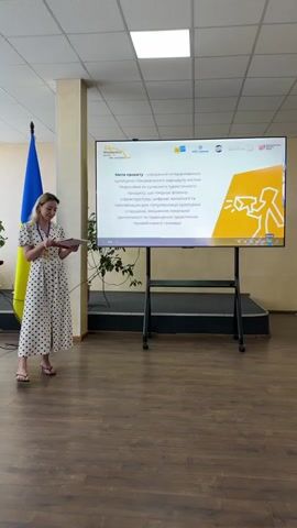 A woman stands to the left of a large screen displaying a presentation. She is wearing a white and black polka dot dress and holding a red folder. She appears to be speaking, possibly presenting the information on the screen. A Ukrainian flag is visible behind her on the left. The screen itself has a title in Ukrainian which translates to "Project Goal: Creation of an interactive cultural and tourist route of the city of Myronivka as a modern tourist product, which combines the physical infrastructure, digital technologies and gamification for popularizing the tourist product and improving the tourist attractiveness of the community." Several logos are also visible on the screen, including "EU", "Creative Europe", and others. The setting appears to be an indoor venue, possibly a conference room or community center, with a wooden floor and a tiled ceiling with fluorescent lighting. The lighting suggests it could be daytime. The woman's expression is neutral and attentive.