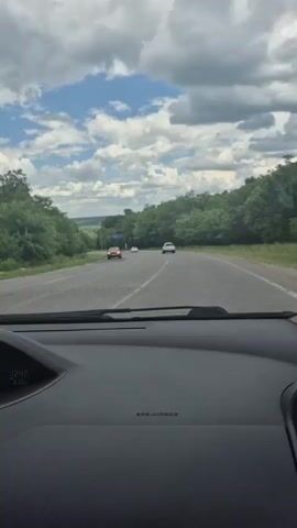 The video shows a view from inside a car driving on a highway. The road is paved and has two lanes in each direction. There are trees and vegetation on both sides of the road, and the sky is cloudy with patches of blue sky visible. There are several other cars on the road, some in front of and some in the opposite lane. The car's dashboard is visible in the foreground, showing the time as 12:40. The video appears to be taken on a sunny day.