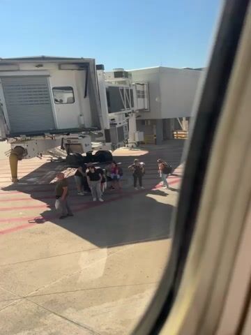 The video shows a group of people standing on the tarmac next to an airplane boarding bridge. The boarding bridge is a large, white, mechanical structure that connects the terminal to the aircraft. The people are of various ages and are wearing casual clothing. Some have luggage with them. The tarmac is made of concrete and has red and white markings. The sky is blue and there are no clouds. The video is taken from inside an airplane looking out through a window.