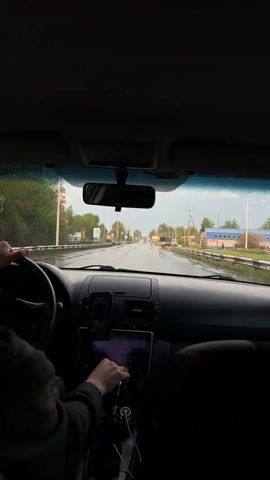 The video shows the interior of a car driving on a wet road, presumably after rain. The dashboard is visible, featuring a phone and a tablet, both displaying screens with images. A person is behind the wheel, their hands occasionally visible as they interact with the tablet. The road ahead is lined with buildings, streetlights, and trees, with a tall industrial chimney visible in the distance. The sky is overcast, and there are raindrops on the windshield.