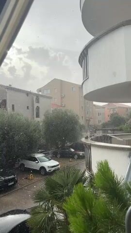 The video shows a view from a window looking out at a residential area during a hailstorm. The sky is cloudy and overcast. Small white pellets, presumably hailstones, are visible falling through the air and accumulating on the ground. In the foreground, green foliage is present. Further back, there are parked cars, including a white SUV, and several multi-story buildings. The hailstorm appears to be active and ongoing.