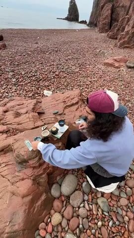 A person with long, curly hair wearing a lavender sweatshirt and a maroon and white baseball cap is seated on a large reddish-brown rock on a pebble beach. They are preparing tea, using a small tea set arranged on the rock. The beach is covered in reddish-brown pebbles, and in the distance there is a tall, jagged rock formation in the water and a tall cliff. The sky is overcast.