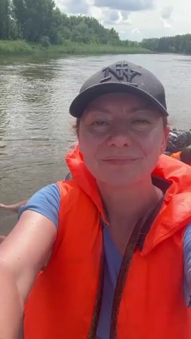 A woman wearing a blue t-shirt and an orange life vest smiles at the camera. She is on a boat with other people. The boat is on a river, with trees on either side of the river. The sky is cloudy. The woman is wearing a black baseball cap with a logo on it. Another person in the boat is wearing a brown hooded jacket. Another person in the boat is wearing a life vest. There are bags and other items in the boat.