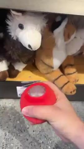 A person is holding and squeezing a red stress ball with a white cross on it. The stress ball has a clear plastic window at the top, through which silvery filling is visible. Behind the hand holding the stress ball are stuffed animals, including a brown and white animal and a tiger. The animals are on a shelf. The floor has a gray speckled pattern.