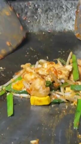 A close-up shot shows food being stir-fried on a griddle. The food consists of pieces of chicken, tofu, bean sprouts, and green onions, all coated in a reddish-orange sauce. A metal spatula is used to toss and stir the ingredients. Some pieces of food are tossed off the griddle and land on the surrounding surface. The griddle appears to be well-used and has some residue from previous cooking. The background is out of focus but suggests an outdoor or food stall environment.