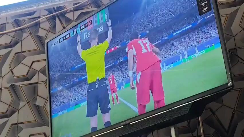 The video shows a television screen displaying a soccer match. A referee in a yellow shirt is holding up a red card to a player in a red jersey. The player is looking down and bending over. Another player in a red jersey runs past them. The score board shows 11:42. The background is a stadium with blurred spectators.
