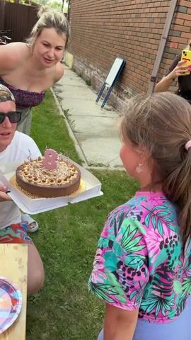 A group of people are gathered outdoors for a birthday celebration. A man in a white t-shirt and a camouflage hat holds a chocolate cake with a lit candle shaped like the number 8. He is smiling at a young girl in a colorful shirt who is clapping her hands. Another woman in a strapless top is also clapping and smiling. A younger child stands to the side. The cake is on a table with decorative paper plates. The environment appears to be a backyard with grass and a brick wall. The overall mood is celebratory.