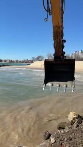 The video shows a construction scene at a beach on a sunny day. A yellow excavator with a black bucket is positioned at the edge of the water. The bucket is lowered towards the water, near a rocky area. In the background, a sandy beach is visible, along with several buildings and a clear blue sky. The water is choppy and appears murky. There are no people visible in the scene.