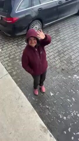 A medium-angle shot shows a person in a hooded, maroon, quilted jacket, black pants, and pink shoes, standing on a gray, tiled surface speckled with moisture and possibly light snow. The person waves and smiles at the camera. To the left of the person is a gray concrete wall. In the background, a black car is parked. The camera angle is looking down on the person from a higher vantage point. The person's body is mostly centered in the frame. After the person waves, the camera angle shifts slightly to the left. A stack of red crates is briefly visible in the background.