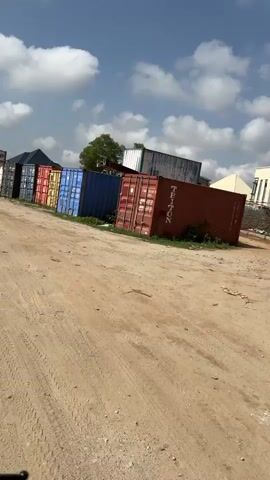 The video shows a row of shipping containers lined up along a dirt road under a partly cloudy sky. A motorcycle with two people on it rides down the dirt road. There are utility poles and a white wall on the right side of the road, and people walking along the road past the wall.