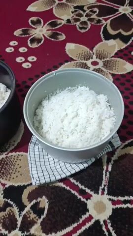 Here is a description of the scene.

The scene features a close-up, static shot of a red and brown floral patterned rug. On the rug are two rice cooker inserts. One is black and contains white rice and a pink spoon. The other is silver and is set on top of a white and black checkered cloth; it also contains white rice.