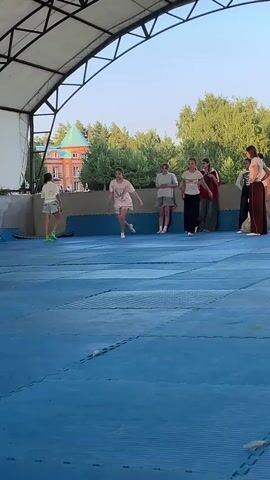 The video shows a group of young people engaged in some sort of physical activity or dance practice on a blue padded floor under a covered outdoor structure. The structure has a white arched roof supported by metal beams. In the background, there is a building with a green roof and trees, suggesting an outdoor recreational area. The sky appears clear, indicating fair weather.

Several individuals are visible, with a few actively participating in what looks like energetic movements, including jumps and squats. One person in the center is mid-air, arms outstretched. Others are observing or preparing to join in. The clothing worn by the participants is casual, typical for a sports or dance setting.

The activity appears to be a group practice or rehearsal, possibly for a performance or a sport. The expressions on their faces are not clearly discernible due to the distance and resolution, but the dynamic movements suggest focus and engagement. The time of day seems to be late afternoon or early evening, given the lighting conditions and the soft shadows. No visible text is present in the video. The setting in Krasnoturansk, Russia, provides a geographical context for the scene.