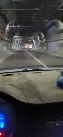 The video is taken from the perspective of someone driving a vehicle through a tunnel. The tunnel appears to be industrial in nature, with exposed pipes and conduits running along the ceiling and walls. The road ahead is dimly lit, with a white line marking the center lane. There are other vehicles visible in the distance, their headlights illuminating the tunnel. The dashboard of the vehicle is visible in the foreground, with a small blue bottle or air freshener placed on it. The lighting in the tunnel creates a hazy effect.