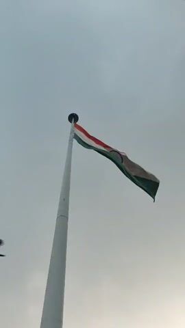 The video opens with a low-angle shot of the Indian flag fluttering on a flagpole against a cloudy sky. The camera then pans down, revealing the base of the flagpole and a railing. In the background, the sun is visible through the clouds, casting a warm glow. Trees and streetlights are also visible, suggesting an outdoor setting like a park or public square. The video continues to pan, showing a glimpse of cars on a road and more trees. The overall mood is peaceful and patriotic.
