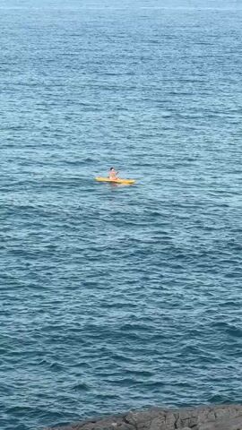 A person is paddleboarding on a calm sea. The person is sitting on a yellow paddleboard and is holding a paddle. The water is blue and there are waves. There are rocks in the foreground of the video.