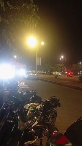 The video shows a street at night, illuminated by streetlights and the headlights of passing vehicles. In the foreground, several motorcycles are parked. In the background, cars and motorcycles are driving on the road. The atmosphere is somewhat blurry due to the low light conditions and the movement of the camera, suggesting it was filmed from a moving vehicle or while walking. The overall impression is of a bustling street scene at night.