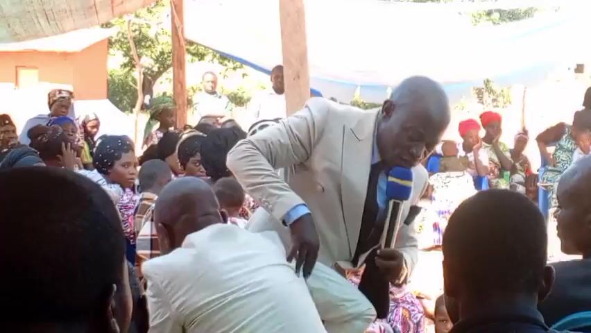 The video captures a man in a light-colored suit, standing outdoors, speaking into a microphone while leaning on the back of a green plastic chair where another man in a light-colored suit is seated. They are surrounded by a gathering of people of various ages, including children and adults, many of whom are looking in their direction. A boy in a white t-shirt is standing to the left of the speaker, looking at a mobile phone. The gathering appears to be under a canopy made of light-colored material, and there's a small building visible in the background. The scene suggests a speech or ceremony taking place within a community setting.