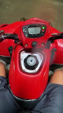 The video shows a first-person view of someone riding a red ATV through a shallow, muddy riverbed in a lush, green forest. The ATV's dashboard is visible, displaying a speed of 0. Ahead, a group of people on ATVs are stopped near a small waterfall cascading down rocks into the river. One individual wearing a green poncho stands near the stopped ATVs. The environment is wet, with water droplets on the camera lens, suggesting rain or spray from the waterfall. The riverbed is narrow, flanked by dense foliage and rocky terrain.