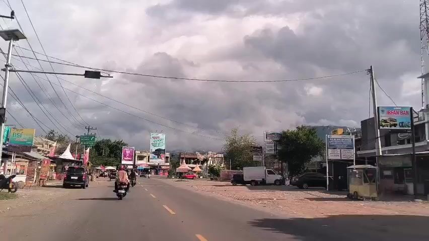 The video depicts a street scene in a town or city. The road, marked with a double yellow center line, is busy with motorbikes and cars. Buildings line both sides of the road, with various commercial signs and billboards visible. Overhead, a network of utility wires crisscrosses the sky. The sky itself is overcast, with heavy, dark clouds suggesting possible rain. The overall impression is of a typical day in a bustling, but somewhat grey, urban setting.